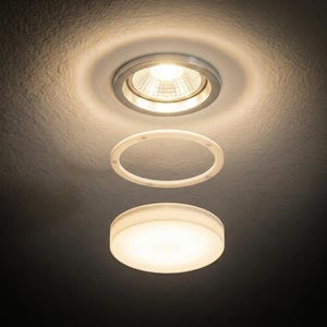 May include: A recessed ceiling light fixture with a bright, focused light. Below it, a white circular ring and a round, glowing light fixture are suspended. The ceiling is textured and painted a light color.