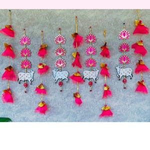 May include: Decorative hanging ornaments with pink lotus flower and cow designs. Each ornament has a pink and gold tassel. The ornaments are suspended from gold strings, creating a festive and colourful display.