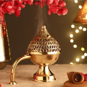 May include: A brass incense burner with a decorative, pierced design. The burner has a handle and a conical lid, with smoke rising from the top. The burner sits on a pedestal base. Red flowers and a small wooden bowl are in the background.