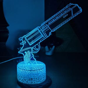 May include: A neon blue gun-shaped lamp. The lamp features a detailed outline of a firearm, illuminated with a bright blue light. The base has a cracked glass design. The lamp is on a dark surface.