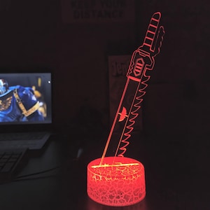 May include: A red LED night light shaped like a futuristic sword with a serrated edge. The base of the light is a textured, red, circular base.