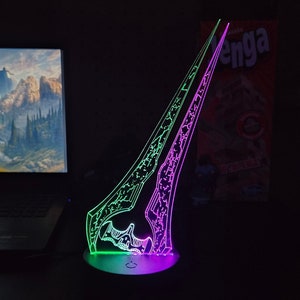 Peut inclure: Une sculpture de lumière LED verte et violette en forme d'épée futuriste, avec une base noire et un interrupteur d'alimentation. La sculpture est éclairée et projette une lueur sur les environs.