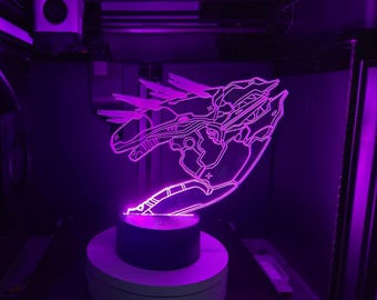 Può includere: Una lampada a LED in acrilico viola che presenta il contorno luminoso e dettagliato di un'astronave o di un veicolo meccanico futuristico. La luce viene emessa da una base nera circolare, illuminando il design inciso con un vibrante bagliore viola neon.