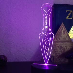 May include: A purple illuminated acrylic knife with the word "QUIETO" printed on it. The knife is on a black base with a purple light shining from the bottom.