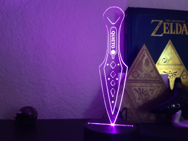 May include: A purple illuminated acrylic knife with the word "QUIETO" printed on it. The knife is on a black base with a purple light shining from the bottom.