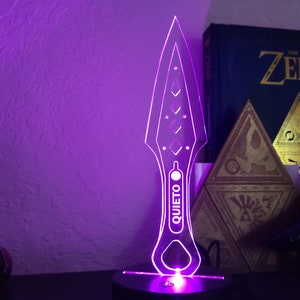 May include: A purple light illuminates a clear acrylic knife with a diamond pattern and the word "QUIETO" printed on the blade. The knife is on a black base.
