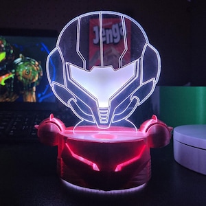 May include: A red and white LED lamp featuring a stylized helmet design. The lamp has a red base with a glowing white outline of the helmet. The word "Jenga" is visible in the background.