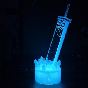 May include: A blue light-up sculpture of a sword with a crystal base. The sword is a replica of the iconic weapon from the Final Fantasy video game series.