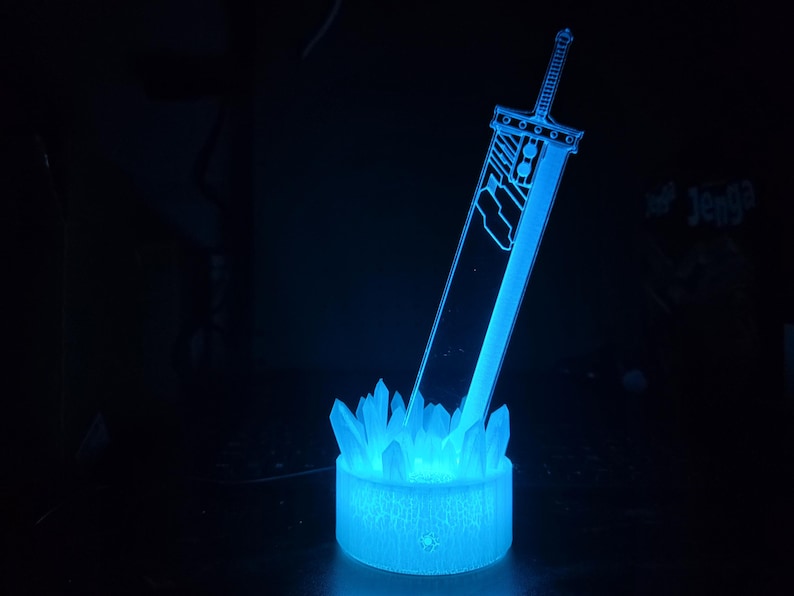 May include: A blue light-up sculpture of a sword with a crystal base. The sword is a replica of the iconic weapon from the Final Fantasy video game series.