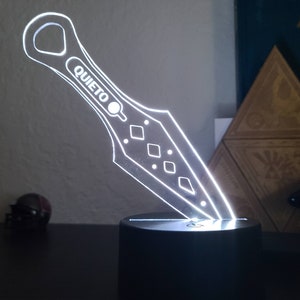 May include: A white acrylic knife-shaped night light with the word "QUIETO" on the handle. The light is on a black base.