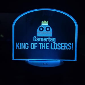 May include: A blue LED light-up sign with the text "Gamertag KING OF THE LOSERS!" and a robot icon wearing a crown. The sign has a rounded top and a base with a power symbol.