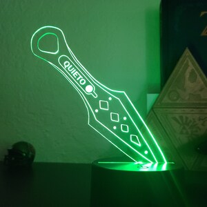 May include: A green illuminated acrylic knife with a diamond pattern and the word "QUIET" on the blade. The knife is on a black base.