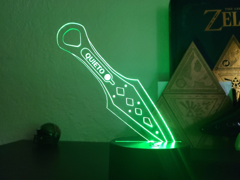 May include: A green illuminated acrylic knife with a diamond pattern and the word "QUIET" on the blade. The knife is on a black base.