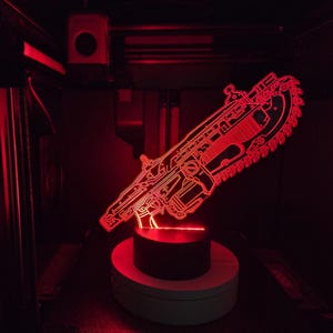 Puede incluir: Una lámpara LED de acrílico rojo brillante con forma de arma de rifle de motosierra. El diseño presenta intrincadas líneas grabadas que se iluminan en rojo brillante, montadas sobre una base negra circular. La luz crea una silueta nítida.