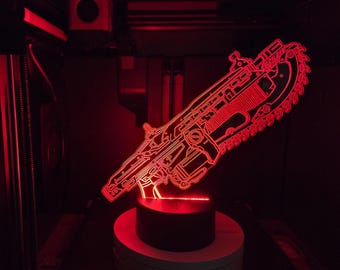 Può includere: Una lampada a LED in acrilico rosso brillante a forma di arma fucile-motosega. Il design presenta intricate linee incise che si illuminano di rosso vivo, montate su una base nera circolare. La luce crea una silhouette nitida e contrastata.