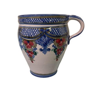 May include: A hand-painted ceramic mug with a blue rim and handle. The mug features a white base with blue and gold decorative patterns, including a lattice design and floral motifs with red and green accents. The mug has a curved handle.