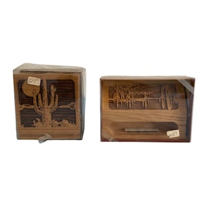 Könnte beinhalten: Zwei Holzkisten mit Wüstenlandschaftsdesigns. Eine Kiste zeigt einen Kaktus und die Sonne, die andere eine Wüstenszene mit Kakteen. Beide sind in klarem Kunststoff eingeschlossen. Auf der ersten Kiste steht die Zahl "1250".