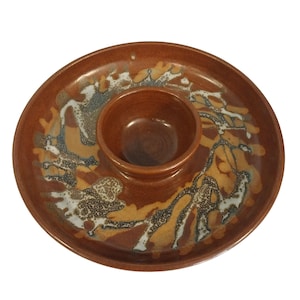 May include: A round, ceramic chip and dip serving dish. The dish is a warm brown color with a central dip bowl. The outer rim features a decorative pattern in shades of brown, yellow, white, and blue.