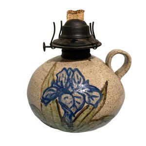 May include: A round, speckled ceramic oil lamp with a beige base and a hand-painted blue iris flower with green leaves. The lamp features a black metal burner assembly at the top with a visible tan wick protruding from the center and a side handle.