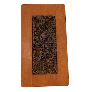 May include: A rectangular wooden plaque with a decorative metal filigree design. The filigree features a floral pattern in a dark bronze color, set within a lighter wood frame. The wood has a warm, natural tone.