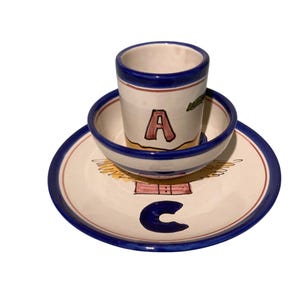 May include: A ceramic set consisting of a plate, a bowl, and a cup. The plate features a blue letter C and a painted wing design. The bowl and cup are decorated with blue rims and the letter A in pink, with additional painted details in green and tan.