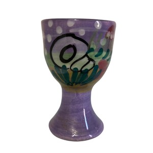 Puede incluir: Un huevero de cerámica pintado a mano con base y cuenco morados. El cuenco presenta un diseño caprichoso con un caracol en blanco y negro, follaje verde y flores rosas y blancas. Lunares blancos adornan el borde superior.