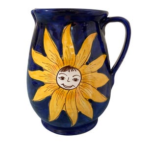 May include: A ceramic pitcher with a dark blue background and a large yellow sunflower design. The sunflower has a smiling face in the center. The pitcher has a handle and a spout.