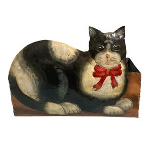 Op de afbeelding: Een metalen houder in de vorm van een kat met een zwart-witte kat en een rode strik op de voorzijde geschilderd. De kat is afgebeeld in een zittende houding met de pootjes ingetrokken op een bruine, rechthoekige basis.