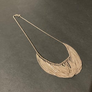 May include: A silver-toned necklace featuring a delicate ball chain that transitions into a multi-layered fringe of fine, dangling metal chains forming a curved, crescent-shaped pendant section that hangs from the main chain.