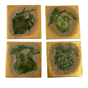 May include: Four square ceramic tiles with a gold background and a raised green fruit design. The fruits include a pear, apple, grapes, and a fruit that looks like a lemon. The tiles are arranged in a square.