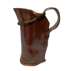 May include: A tall, hand-thrown ceramic jug with a glossy, deep reddish-brown glaze and a speckled finish. The vessel features an asymmetrical, wavy rim, a curved handle, and a decorative, twisted rope-like clay accent encircling the upper neck.