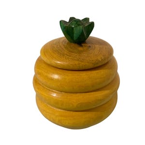 May include: A wooden decorative box with a lid and a green floral-shaped handle. The box is made of stacked, rounded layers of light brown wood. The lid is also made of wood and has a circular shape.
