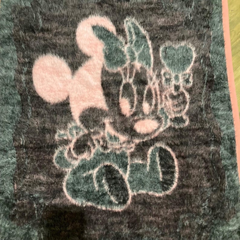 May include: A rectangular fuzzy rug featuring a dark gray background with a light pink graphic of a cartoon mouse character wearing a bow. The character is depicted in a seated position with a heart-shaped icon positioned to the right side.