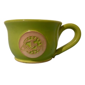 May include: A lime green ceramic mug with a rounded handle. The mug features a circular, tan-colored emblem with the word "Love" in a stylized font. The mug's interior and handle match the exterior color.
