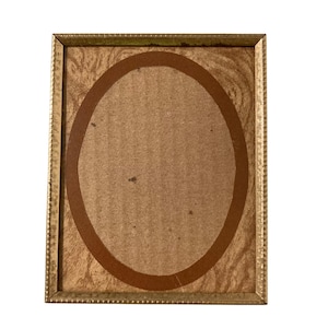 May include: A rectangular picture frame with a metallic gold finish and a textured, beaded edge. The frame contains a light brown, wood-grain patterned backing paper featuring a large, centred dark brown oval shape printed on the surface.