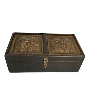 May include: A rectangular wooden box with a dark finish. The lid features two brass panels with ornate floral designs. A small brass clasp secures the box, which is likely used for storage or display.