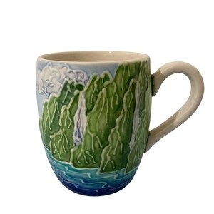 Puede incluir: Una taza de cerámica con un diseño en relieve texturizado de montañas verdes y cascadas blancas sobre un cielo azul claro. La base de la taza muestra olas de agua azules estilizadas. Tiene un asa y un interior de color crema suave.
