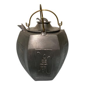 May include: A metallic gray hexagonal teapot with a brushed finish, featuring a brass-colored arched handle and a lid topped with a small animal figurine. The body displays embossed rectangular panels with intricate Asian-style characters.