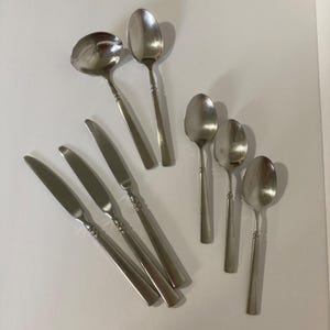 Op de afbeelding: Een verzameling roestvrijstalen bestek, waaronder lepels en messen. De set bevat een soeplepel, verschillende lepels van verschillende maten en meerdere messen. Het bestek heeft een eenvoudig, elegant ontwerp met een gepolijste afwerking.