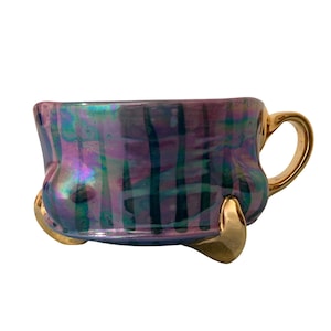 May include: Iridescent teacup with a gold handle and three gold feet. The cup has a purple, blue, and green plaid pattern. The cup is a unique and decorative item.