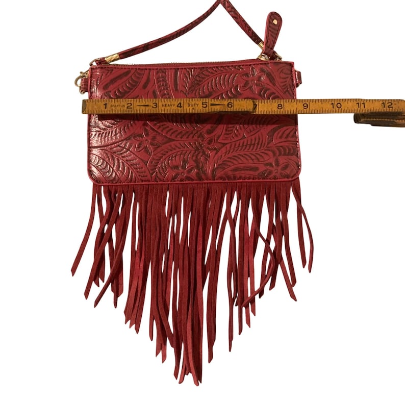 Puede incluir: Bolso bandolera rojo con dise&ntilde;o floral en relieve y flecos. El bolso mide aproximadamente 20 cm de ancho. Cuenta con un asa superior y una correa de hombro desmontable. El bolso tiene cierre de cremallera y herrajes dorados.