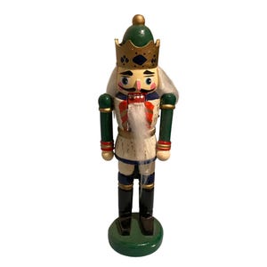 May include: A wooden nutcracker figurine wearing a green and gold crown, a green jacket with red and gold trim, and black boots. It features a white faux fur beard, painted facial details, and stands on a circular green base. The design includes blue accents on the crown and uniform, with a white faux fur hair element.