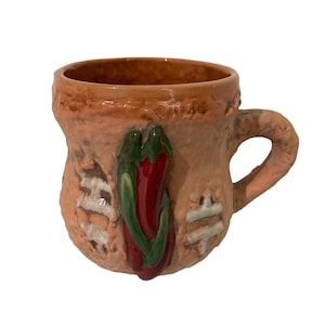 Könnte beinhalten: Handgefertigte Keramiktasse mit strukturierter, pfirsichfarbener Außenseite. Eine rote Chilischote mit grünen Stielen ist an der Seite befestigt. Die Tasse hat einen abgerundeten Henkel und ein rustikales Aussehen, was auf Handarbeit hindeutet.