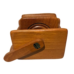 May include: A set of square wooden coasters stored in a matching rectangular wooden holder. The light brown wood features a visible grain pattern. A small, dark rectangular label with the word dolphin is attached to the front of the holder.