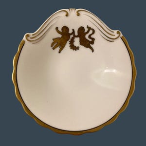 Puede incluir: Un plato blanco con borde festoneado, borde dorado y detalles decorativos. En el centro se representan dos figuras de querubines dorados, sosteniendo una corona. El plato tiene un acabado brillante y un diseño clásico.