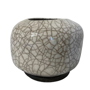 May include: A round, off-white ceramic vase with a cracked glaze pattern. The vase has a dark brown base and a wide opening at the top. The surface is covered in a network of fine, dark lines.