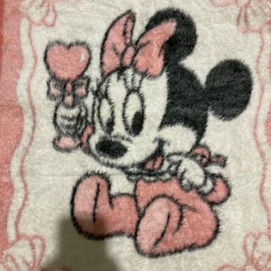 May include: A fuzzy white and pink blanket featuring a graphic of Minnie Mouse wearing a pink bow, holding a pink heart-shaped wand. The design is framed by a light pink border with bow accents at the corners, rendered in a soft, textured material.
