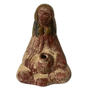 May include: A terracotta-colored ceramic sculpture of a seated figure with a textured, mottled surface. The figure wears a smooth, metallic-toned headpiece and holds a small, circular vessel at the center of the lap. The overall form is conical.