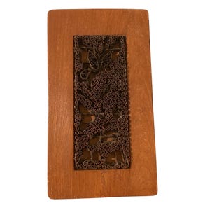 May include: A rectangular wooden block featuring a central inset panel of dark metal wirework. The metal insert displays a dense, intricate pattern of interconnected swirls and loops, contrasting with the smooth, light brown wood frame.