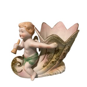 May include: A porcelain figurine of a cherub with blonde hair holding a small trumpet, seated on a shell-shaped vessel. The cherub wears a green cloth, and the vessel features a scalloped rim with pink accents and gold-toned decorative detailing.